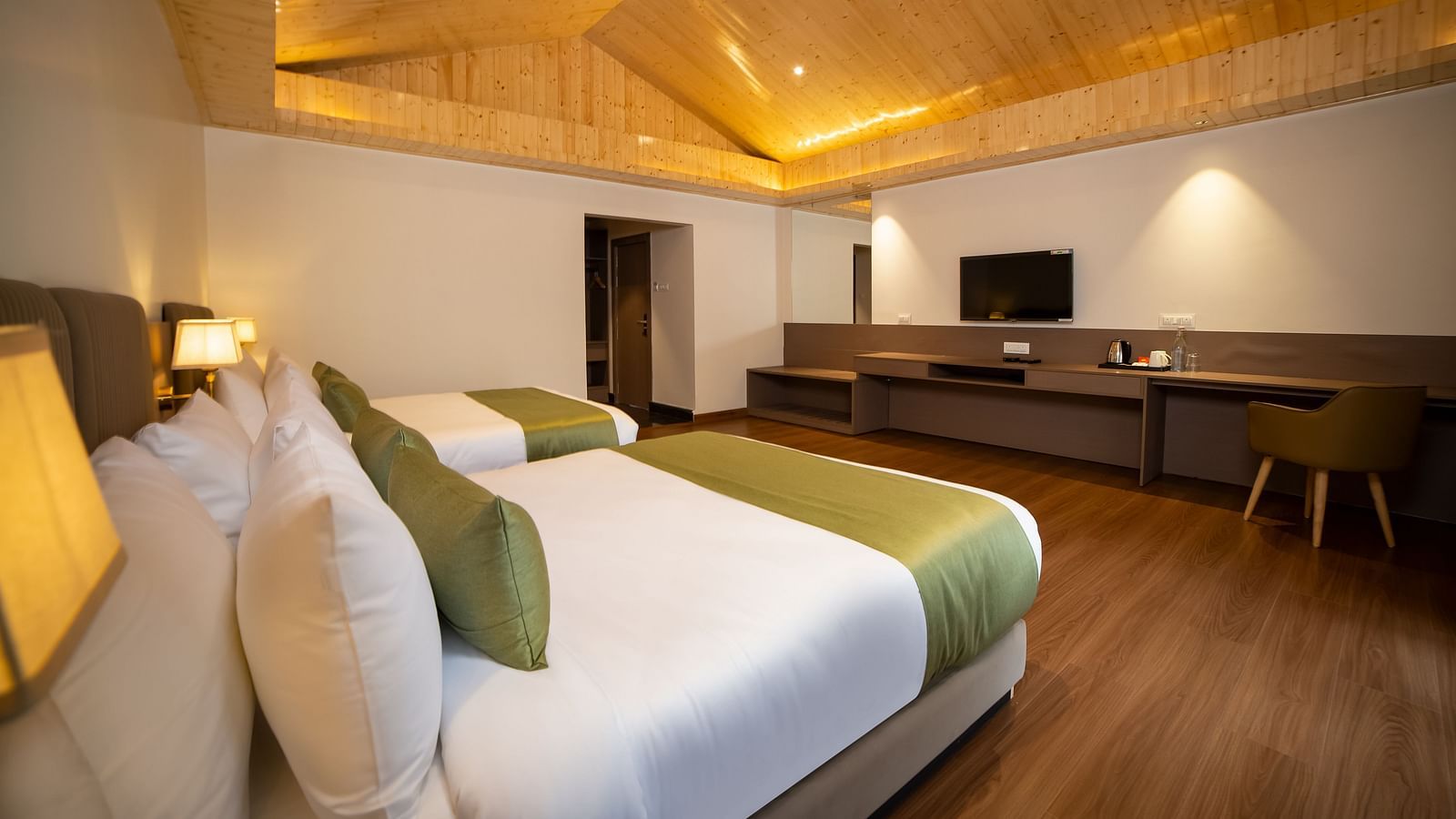 Modern hotel room with two neatly made beds featuring green accents, wooden flooring, a long desk with a chair, a wall-mounted TV, and warm ambient ceiling lighting - Yashshree Pelling Chekhim Retreat.