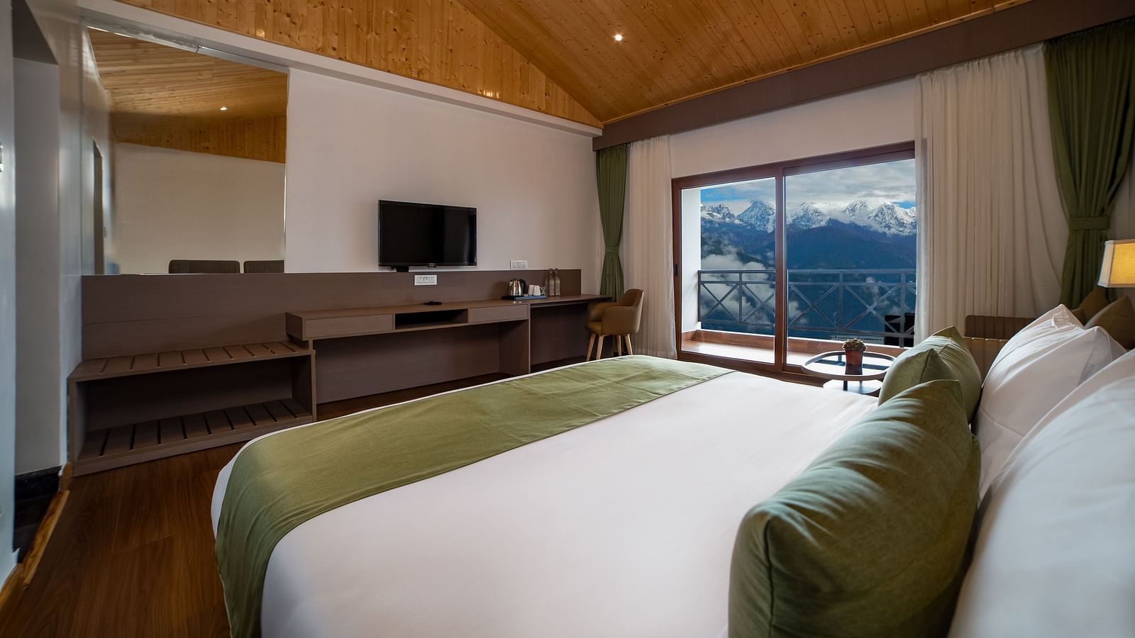 Hotel room interior featuring a neatly made bed with green accents, wooden ceiling, a wall-mounted TV with a desk, and a large window offering a scenic mountain view - Yashshree Pelling Chekhim Retreat.