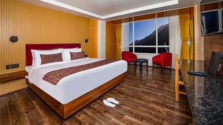 Well-appointed hotel room with a large bed, red headboard, patterned bedding, polished wooden floors, red accent chairs by a large window with mountain views, and a work desk with a mounted TV - Yashshree Lachen.