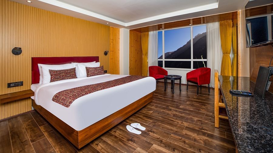 Well-appointed hotel room with a large bed, red headboard, patterned bedding, polished wooden floors, red accent chairs by a large window with mountain views, and a work desk with a mounted TV - Yashshree Lachen.