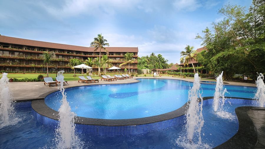 a large, circular swimming pool, water fountains, sun loungers, palm trees, and a tropical ambience under a blue sky at Uday Backwater Resort