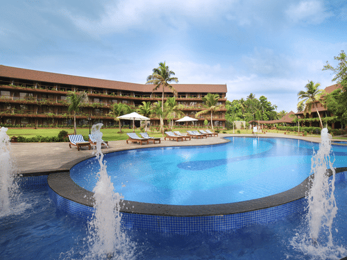 a large, circular swimming pool, water fountains, sun loungers, palm trees, and a tropical ambience under a blue sky at Uday Backwater Resort