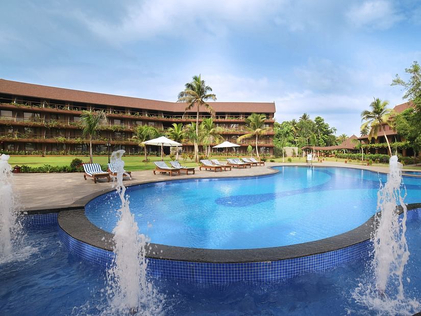 a large, circular swimming pool, water fountains, sun loungers, palm trees, and a tropical ambience under a blue sky at Uday Backwater Resort