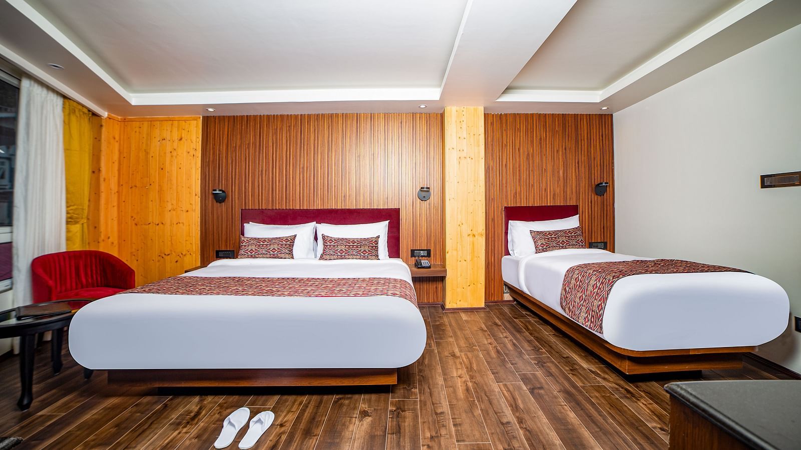  Bright and modern hotel room with wooden flooring, a double bed and a single bed both featuring white linens and red headboards, a pair of white slippers on the floor, and soft lighting accenting the wood-panelled walls - Yashshree Lachen.