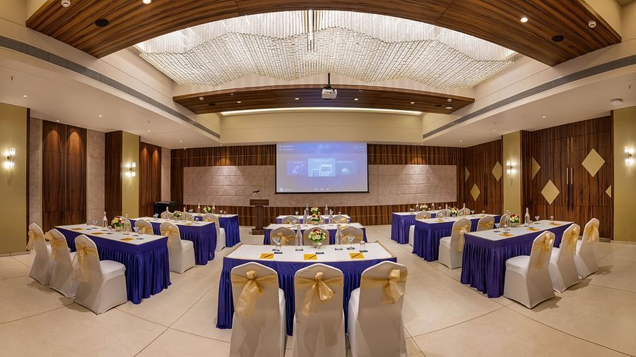 Banquet hall featuring a round-table setup, high ceilings and a projector screentowering hotel buildings featuring balconies, a signage with hotel name, and surrounded by lush greenery, set against a cloudy sky - Golden Tulip, Candolim, Goa