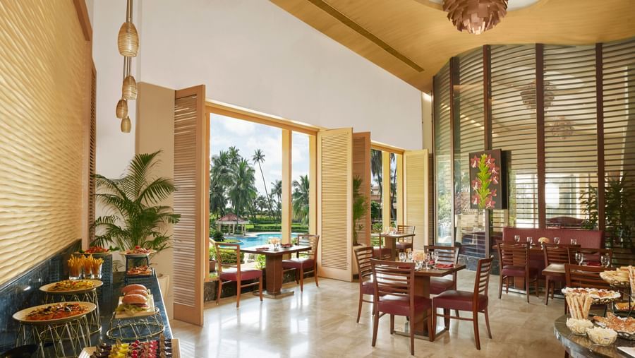 An indoor dining area with wooden tables and chairs near large windows overlooking greenery at The Retreat Hotel & Convention Centre, one of the hotels with membership, Mumbai.