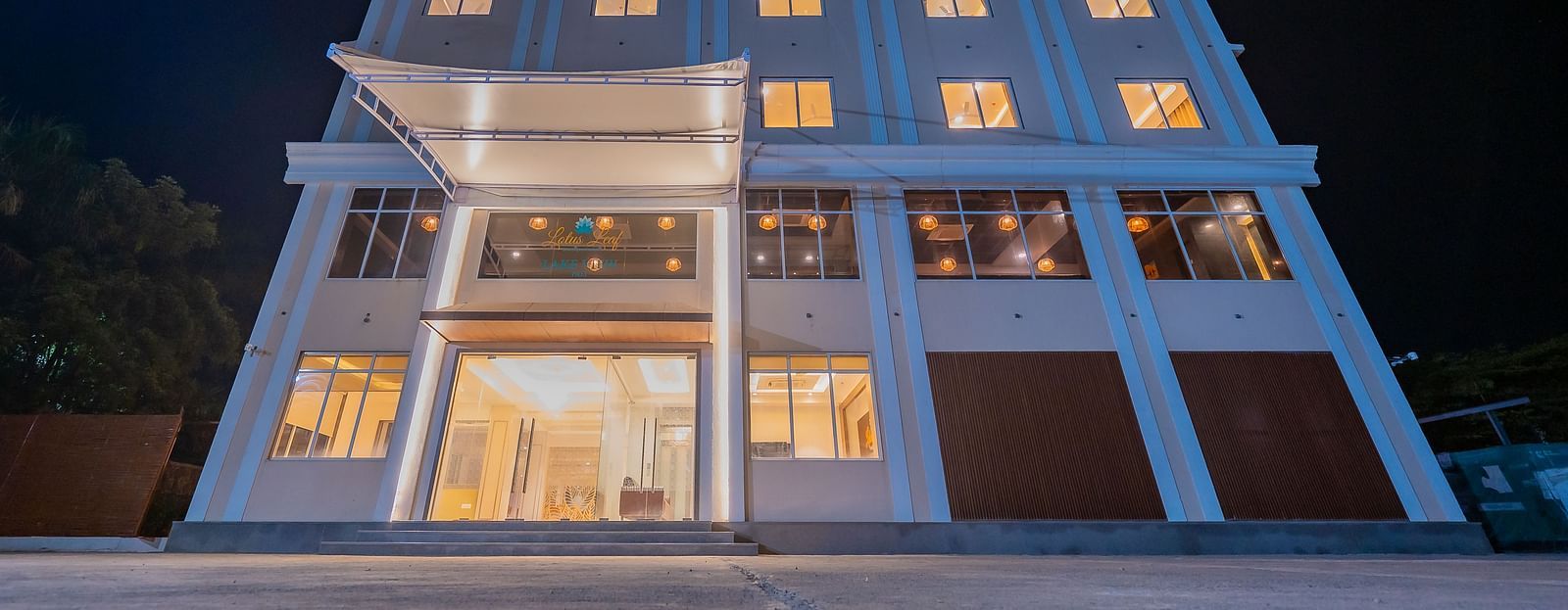 A night view of a modern multi-storey hotel building, brightly lit with warm lights emanating from the windows and entrance at Lotus Leaf Boutique Hotel.