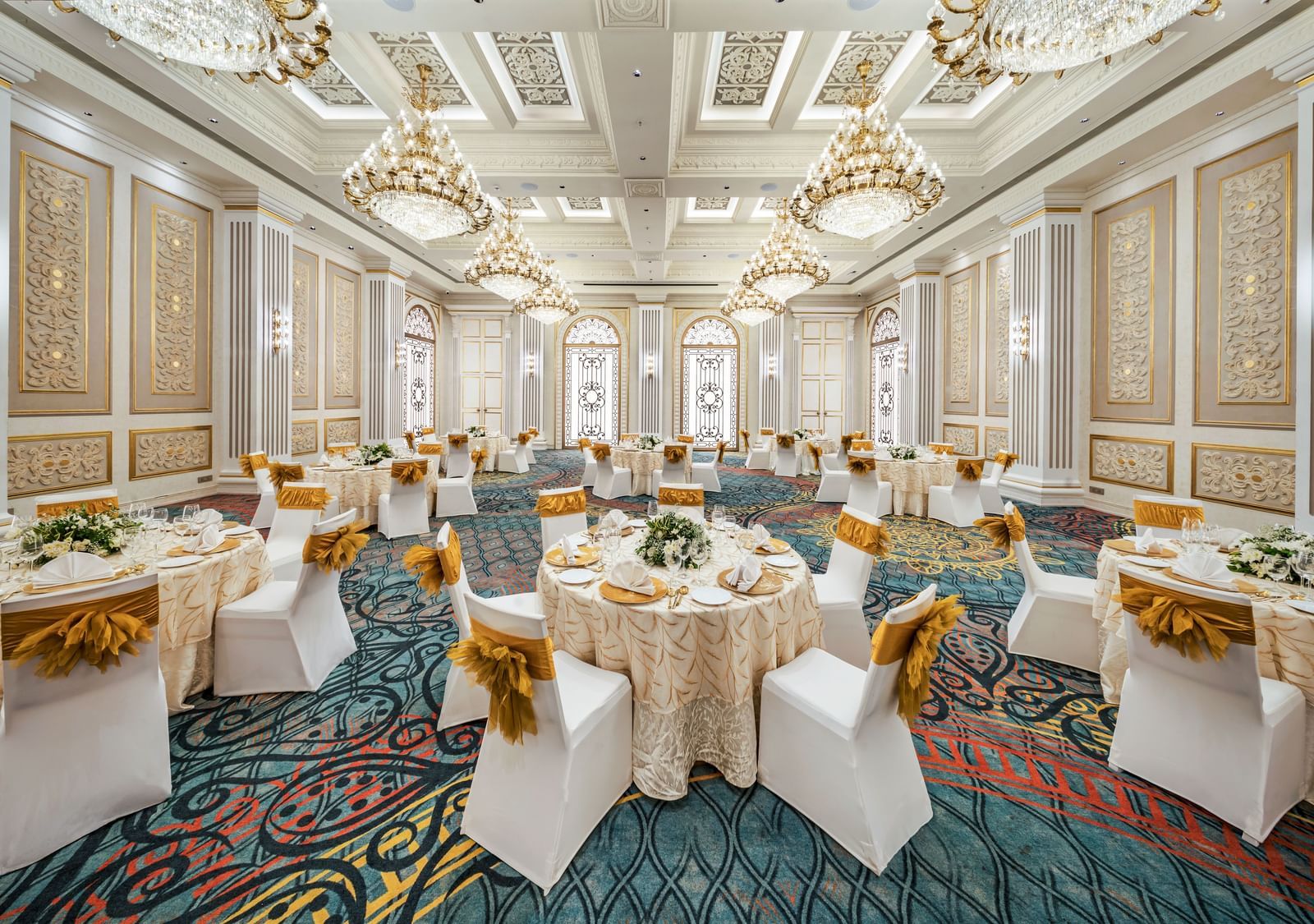 A grand hall prepared for a reception, showcasing elegant round tables with golden accents and white chairs, all beneath a ceiling adorned with multiple sparkling chandeliers.