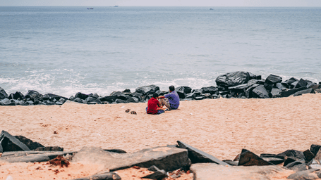 A sandy and rocky beach with the ocean in the foreground and a person in red sitting near the rocks