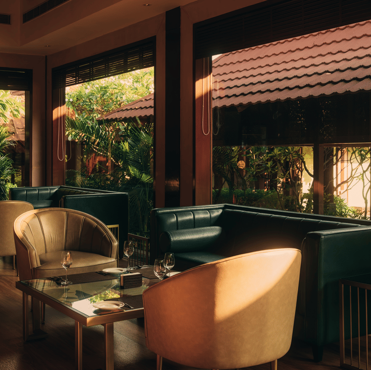 A lounge area at The Ananta Udaipur, featuring comfortable seating, wooden furniture, and  overlooking the lush greenery.