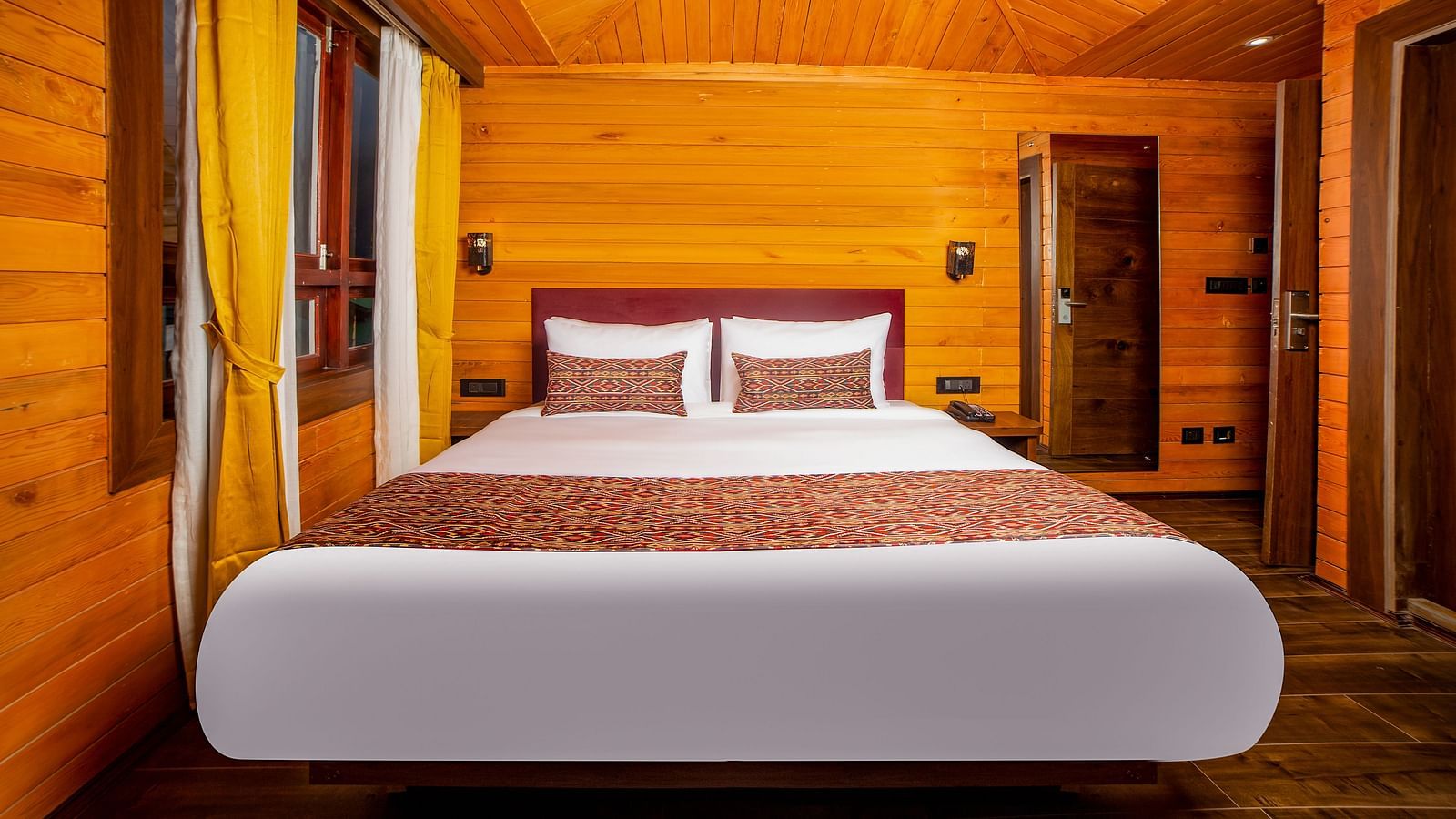 A hotel room with wooden walls and ceiling, featuring a large bed with crisp white linens and red-patterned accents, two windows with yellow curtains, and a wooden door and closet in the background - Yashshree Lachen.