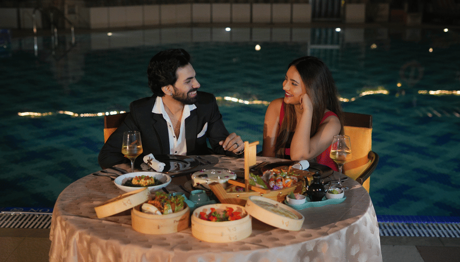 A man and a woman seated at a round table beside a swimming pool with plated food and wine glasses at The Suryaa, New Delhi.