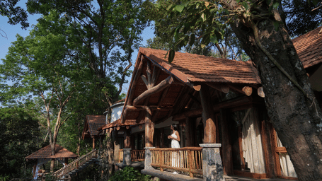 Veranda at Estate Paathiri with a tiled gable roof on log posts with stone bases, a pole balustrade and tall trees around at Earthitects Holiday Experiences.