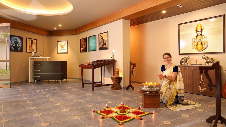 A woman in traditional attire sits beside a beautifully decorated rangoli with oil lamps, in an elegant, warmly lit cultural space at Uday Backwater Resort
