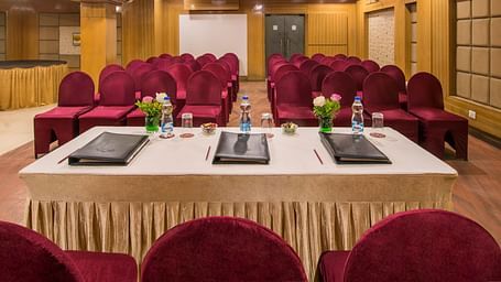 A wooden furnished boardroom at Lords Eco Inn Dahej, featuring a panel table set facing the rows of arranged red chairs in the background.