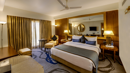  A well-appointed hotel room featuring a large bed with a teal throw, a patterned rug, a seating area, dark gold curtains, and warm lighting from lamps and overhead fixtures - Pride Elite Daman