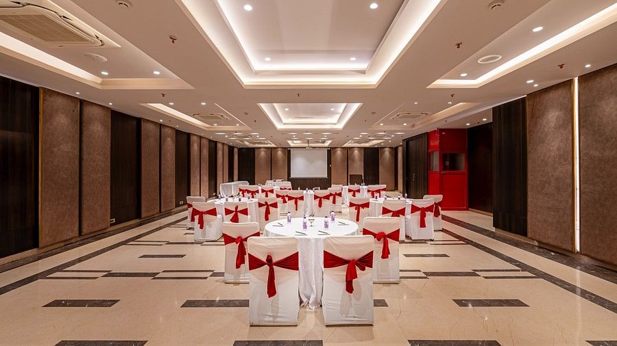 This conference hall is arranged with a projector screen at one end, and round tables surrounded by white-covered chairs, each adorned with a red ribbon.