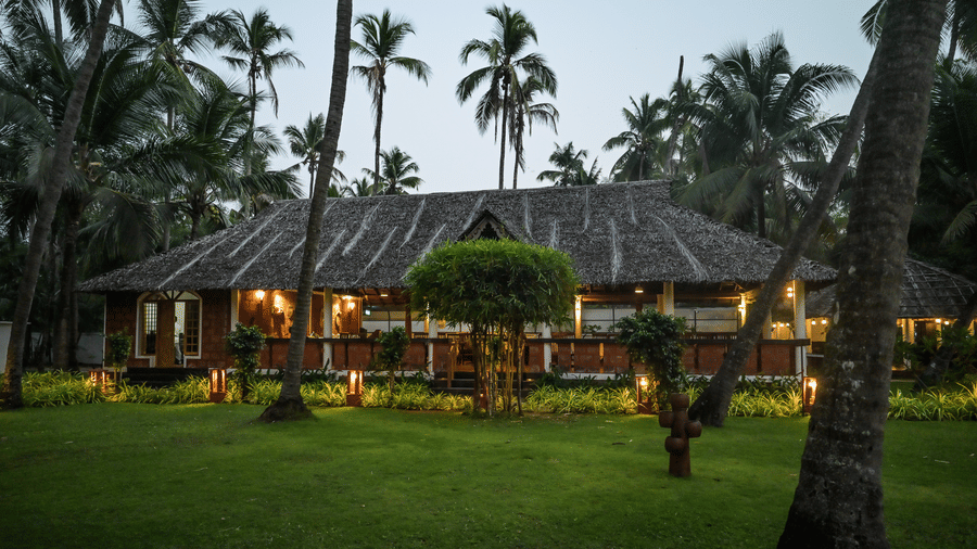 Dusk exterior view of Ayur Villas at Ayur on the Beach Nattika featuring warmly lit traditional tiled roof cottages surrounded by tall coconut palm trees and green lawns.