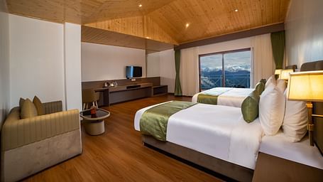 Spacious modern hotel room with two neatly made twin beds, green accents, a wooden floor and ceiling, a seating area with a sofa and table, a flat-screen TV, and large windows offering a scenic mountain view - Yashshree Pelling Chekhim Retreat.