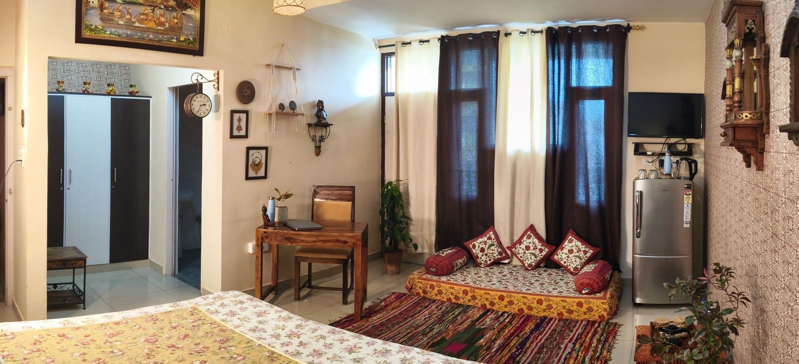 A room with a bed, cushioned seating near the window, desk and wall-mounted items at Jaypore360 Homestay in Jaipur.
