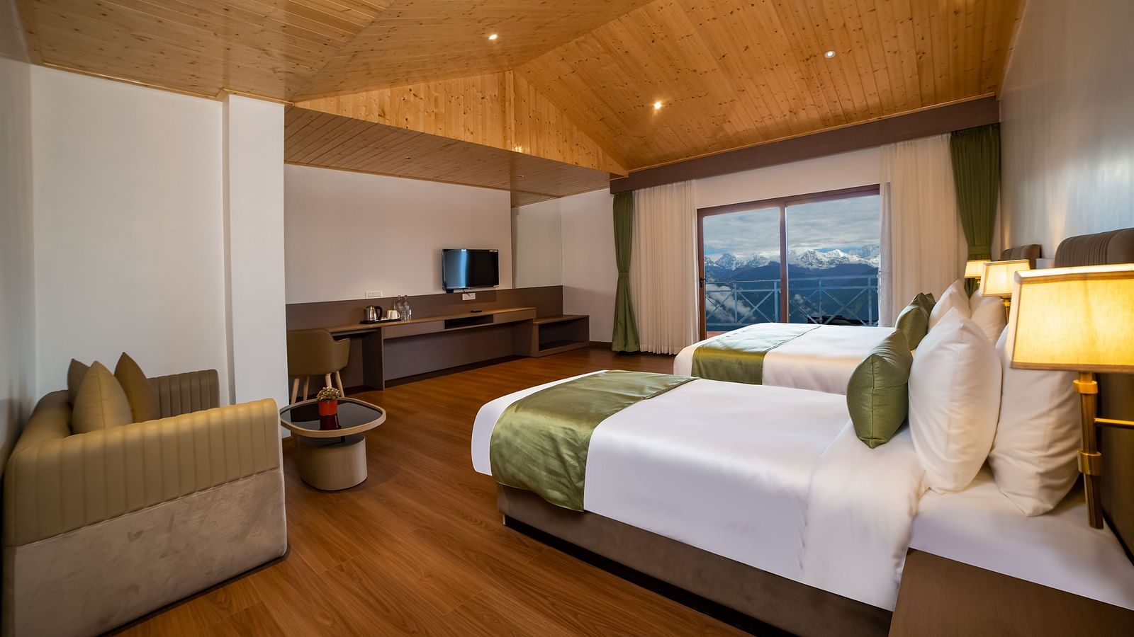 Spacious modern hotel room with two neatly made twin beds, green accents, a wooden floor and ceiling, a seating area with a sofa and table, a flat-screen TV, and large windows offering a scenic mountain view - Yashshree Pelling Chekhim Retreat.