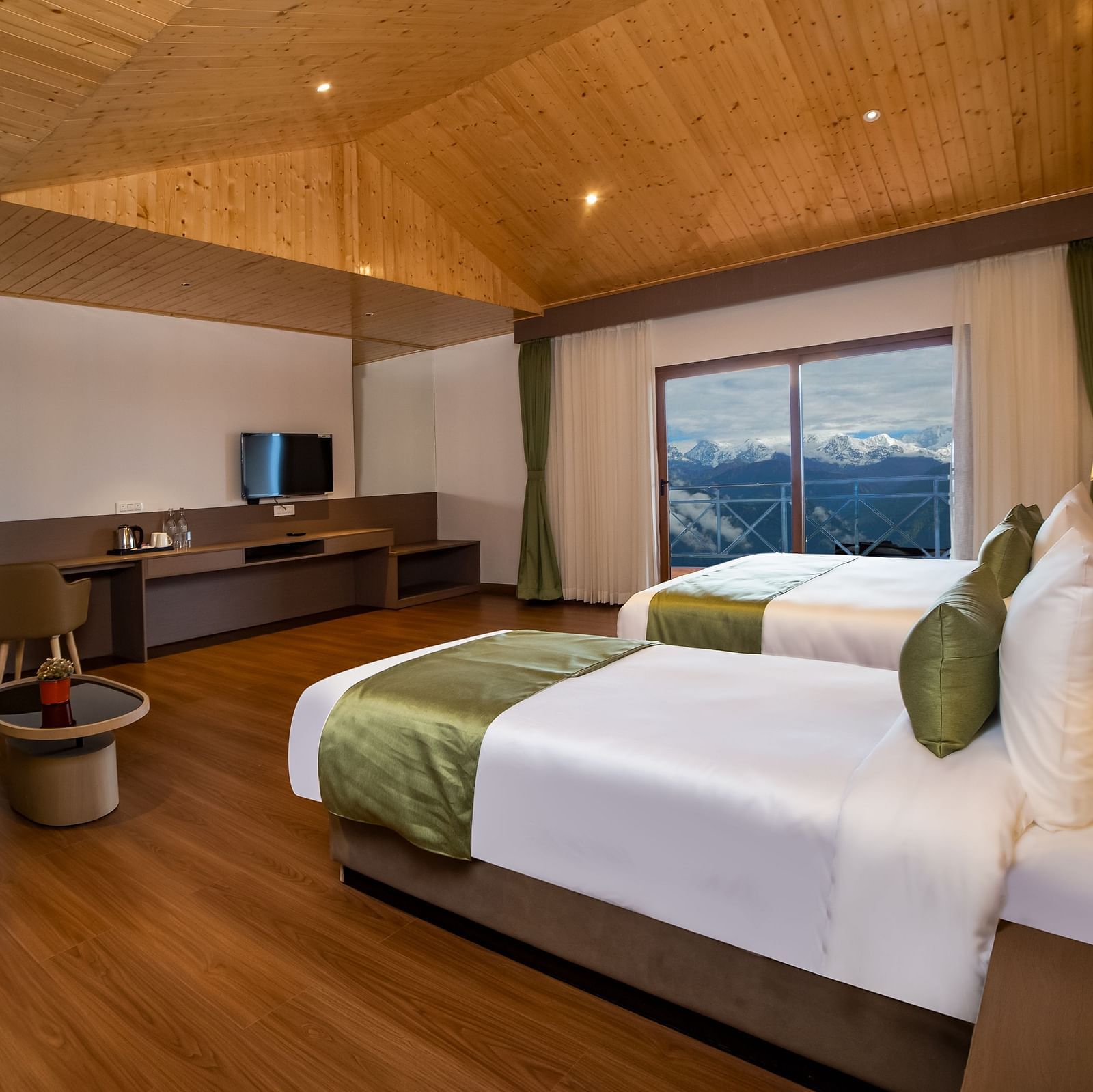 Spacious modern hotel room with two neatly made twin beds, green accents, a wooden floor and ceiling, a seating area with a sofa and table, a flat-screen TV, and large windows offering a scenic mountain view - Yashshree Pelling Chekhim Retreat.