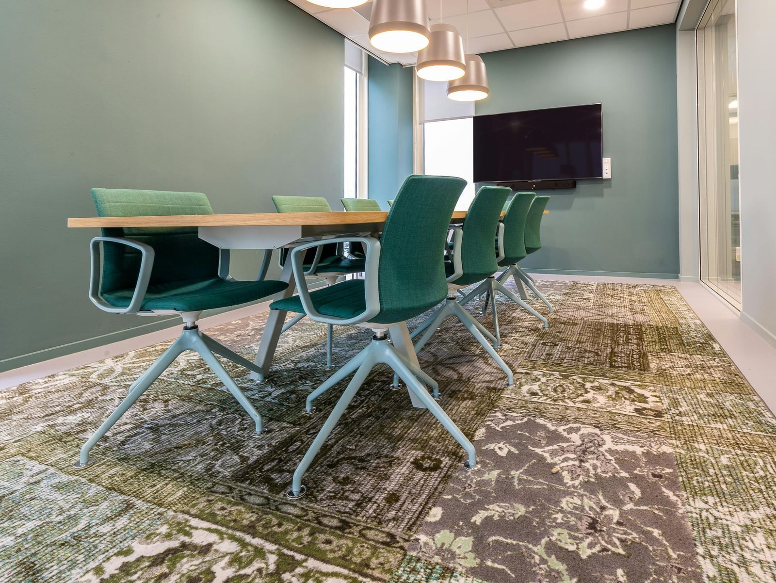 Modern meeting room with green chairs around a wooden table, a large screen on the wall, and patterned carpet flooring.