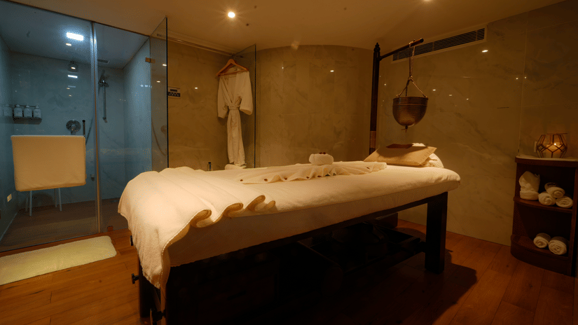 Massage room at The Suryaa New Delhi with warm lighting, a massage table with white linens, a glass-enclosed shower area, a hanging bathrobe