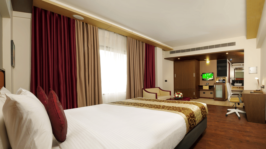 The Deluxe Room at Dimora, Thiruvananthapuram with a king size bed, a large glass window with curtains, an armchair, a work desk, and wooden flooring.