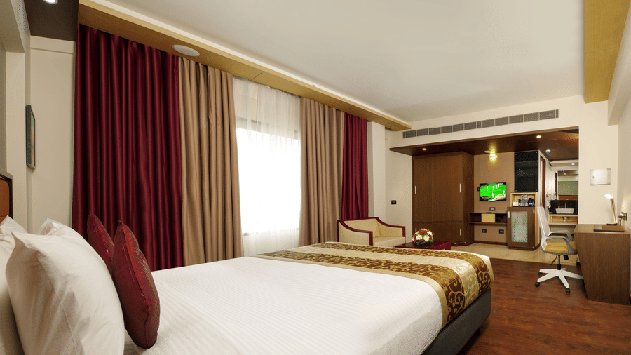 The Deluxe Room at Dimora, Thiruvananthapuram with a king size bed, a large glass window with curtains, an armchair, a work desk, and wooden flooring.