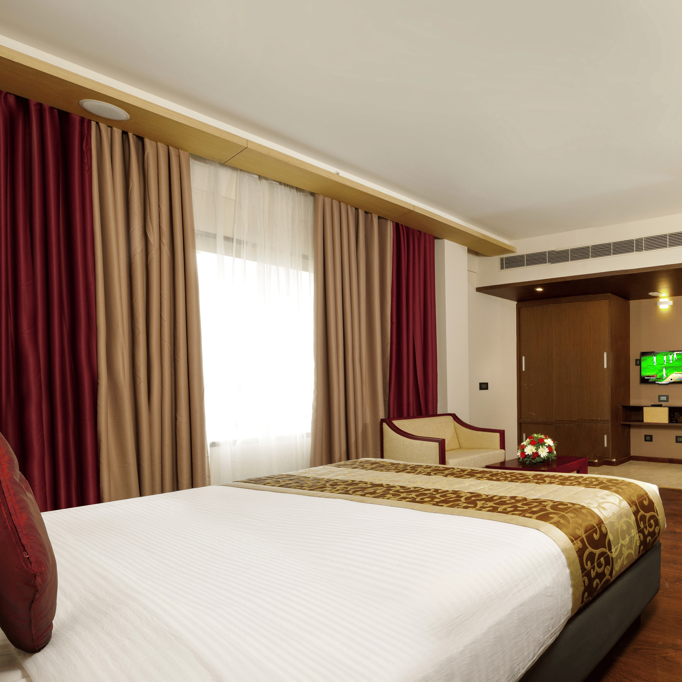 The Deluxe Room at Dimora, Thiruvananthapuram with a king size bed, a large glass window with curtains, an armchair, a work desk, and wooden flooring.