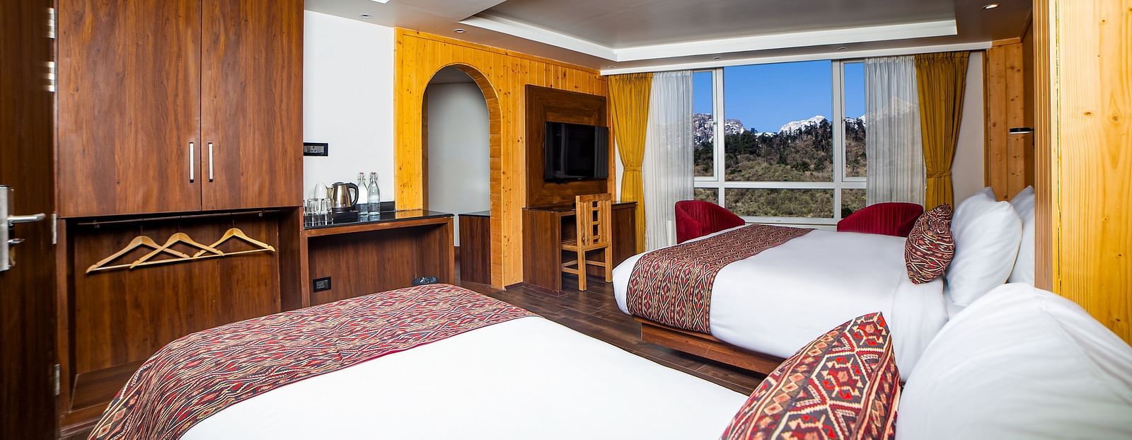  Spacious hotel room featuring two double beds with red and patterned accents, wooden furnishings, a flat-screen TV, large windows offering a scenic view, and a wardrobe with an open hanging area - Yashshree Lachen.