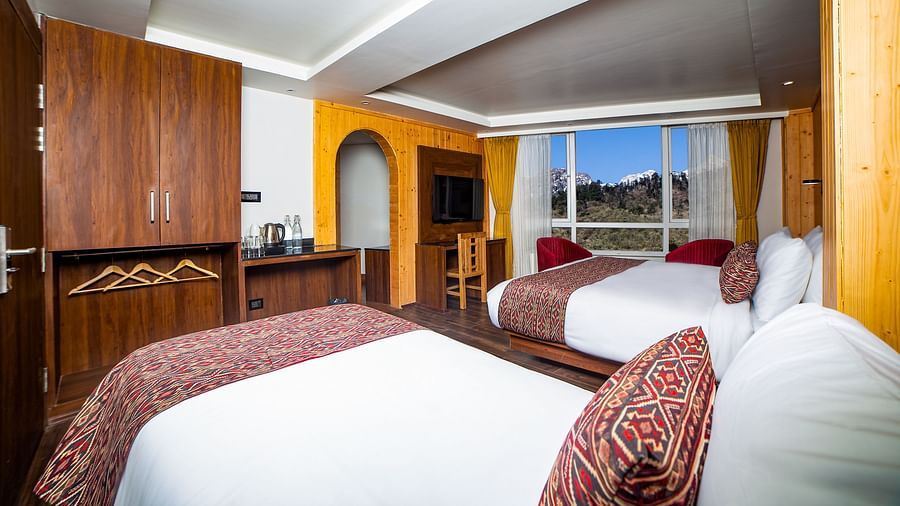  Spacious hotel room featuring two double beds with red and patterned accents, wooden furnishings, a flat-screen TV, large windows offering a scenic view, and a wardrobe with an open hanging area - Yashshree Lachen.