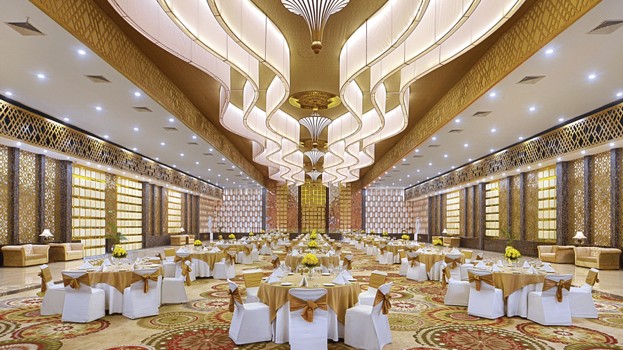 A grand banquet hall at The Bristol Hotel, Gurgaon set with round tables, white chair covers with gold sashes, and an ornate sculptural ceiling.