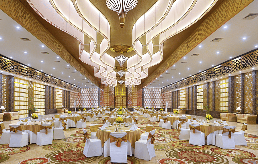 A grand banquet hall at The Bristol Hotel, Gurgaon set with round tables, white chair covers with gold sashes, and an ornate sculptural ceiling.