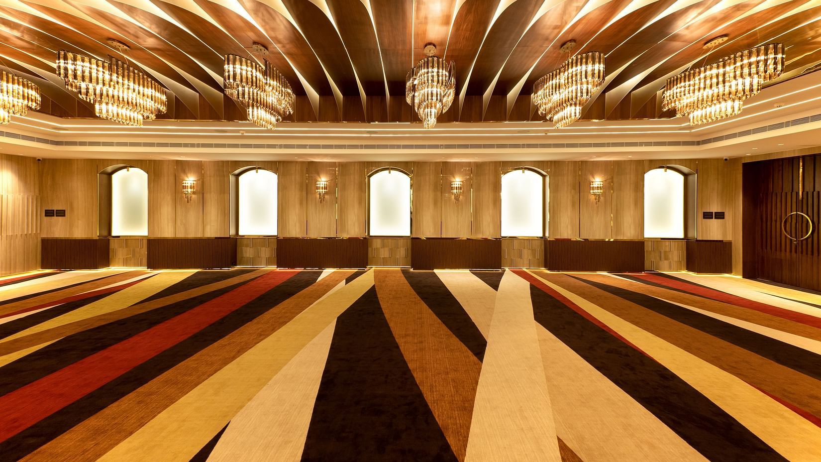 Striped carpeted hall with arched frosted windows, gold chandeliers and curved wooden ceiling panels at Jenneys Residency, Coimbatore