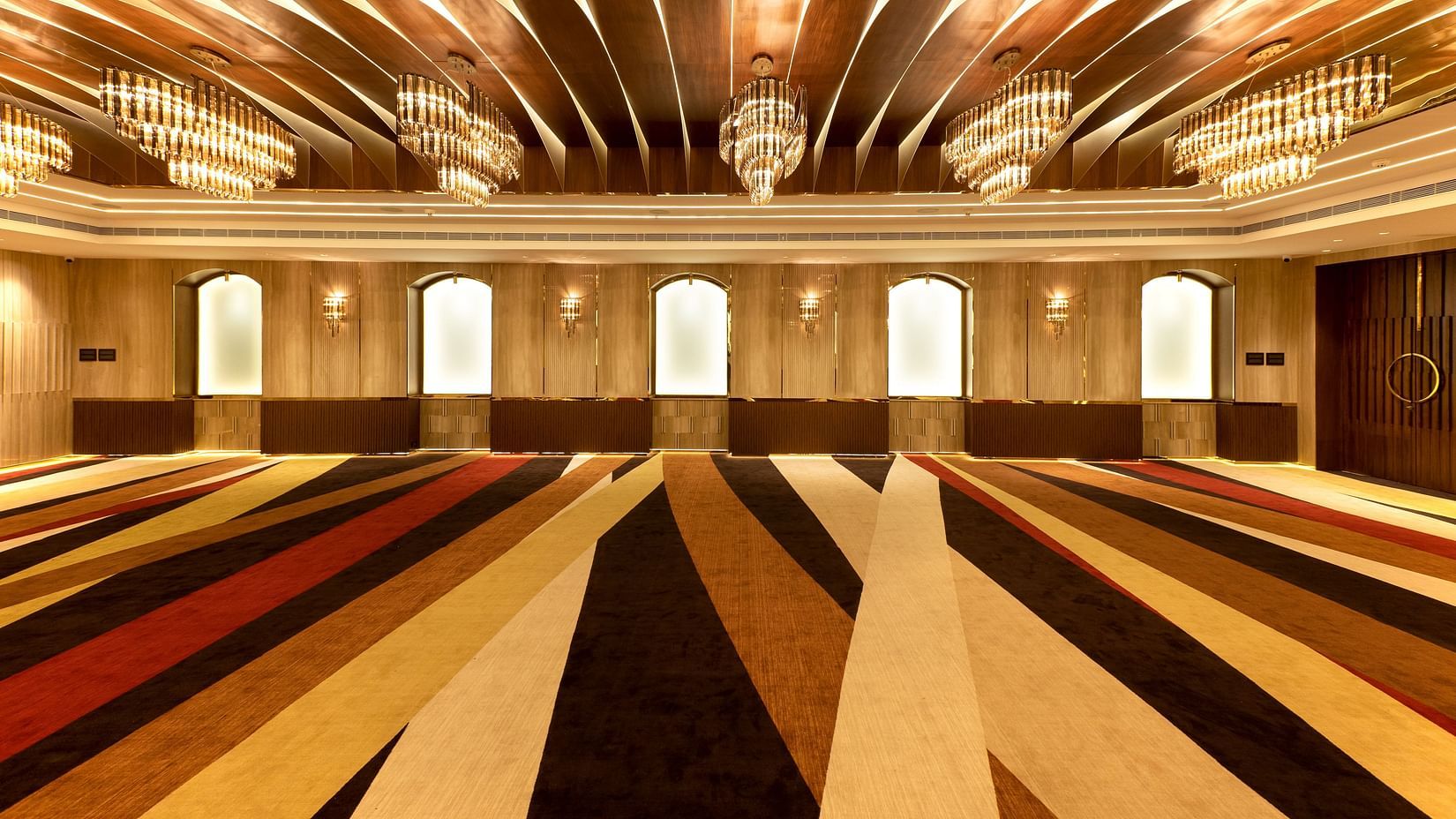 Striped carpeted hall with arched frosted windows, gold chandeliers and curved wooden ceiling panels - Jenneys Residency, Coimbatore