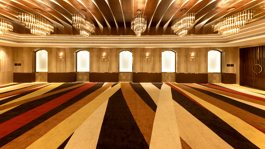 Striped carpeted hall with arched frosted windows, gold chandeliers and curved wooden ceiling panels at Jenneys Residency, Coimbatore