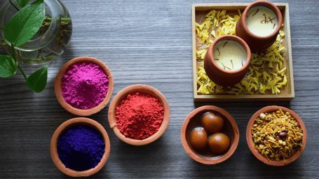 A flat-lay of traditional Holi treats, including bowls of coloured powder, sweet gulab jamun, and glasses of thandai on a wooden surface.