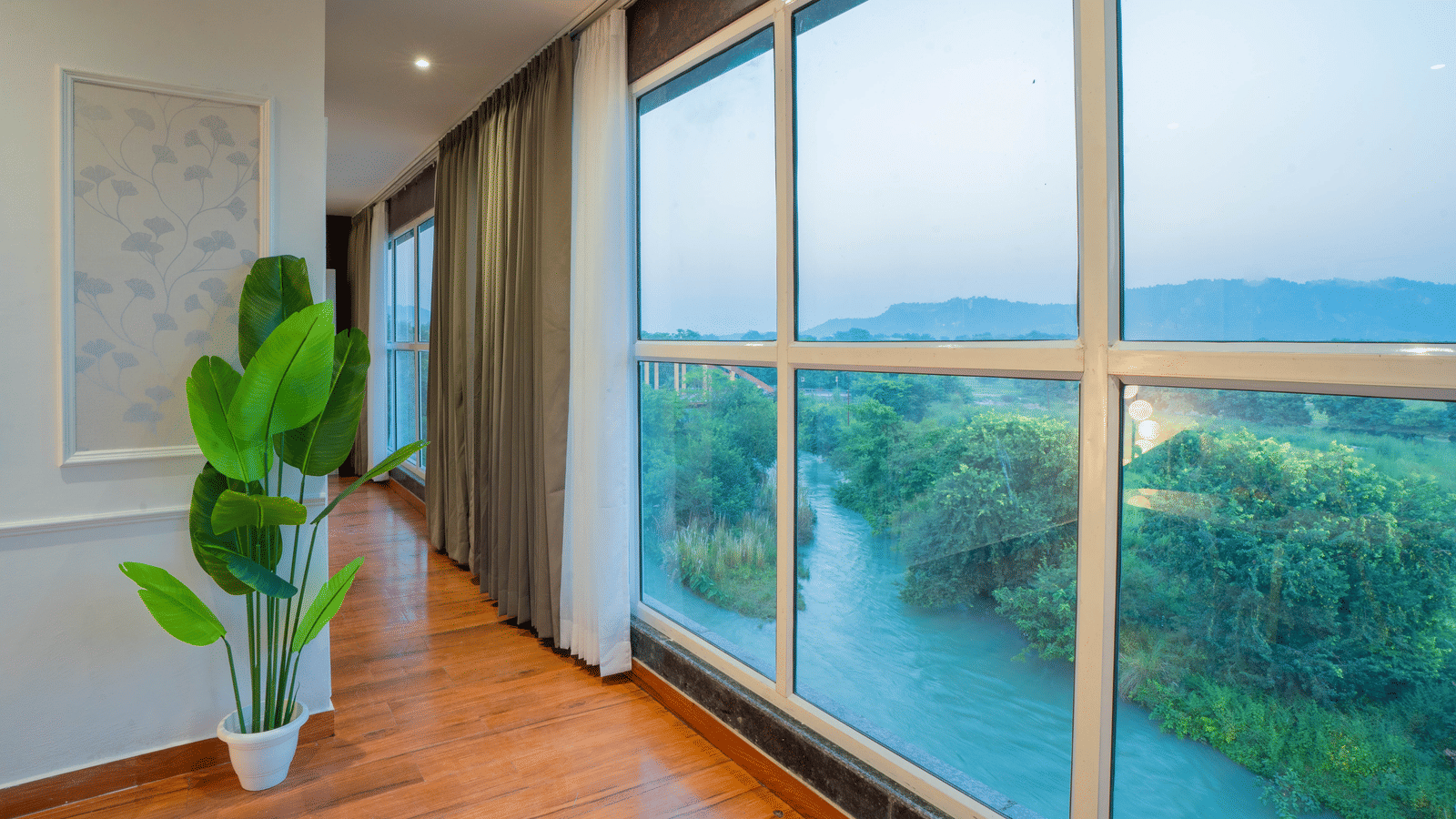 A hallway at Sukoon by the Ganges features floor-to-ceiling windows offering a tranquil view of the river and green landscape.