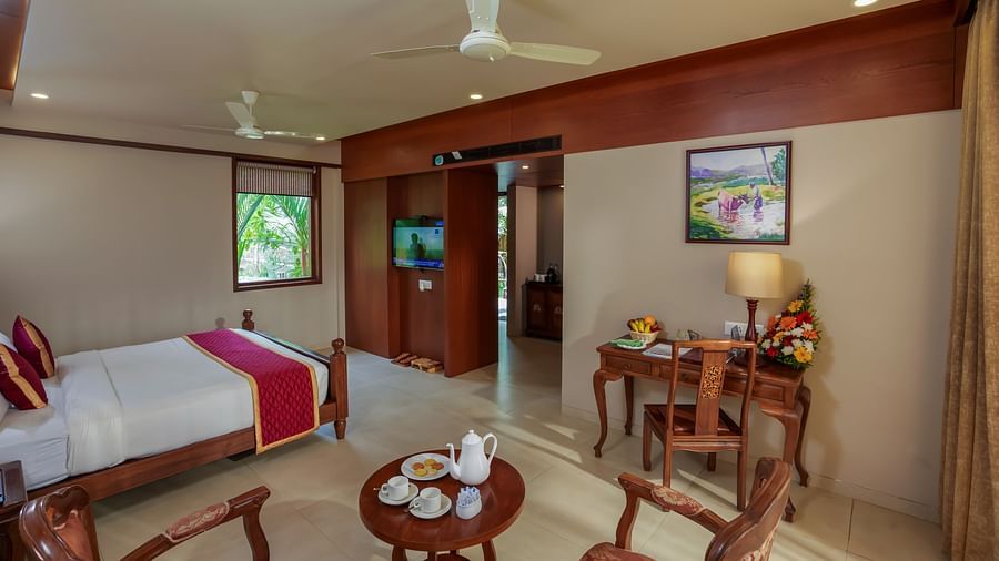 A room at Uday Backwater Resort featuring a plush bed faing the TV on the wall with a small window next to it and a work desk on the other side of the room with sitting space with high tea served on the coffee table