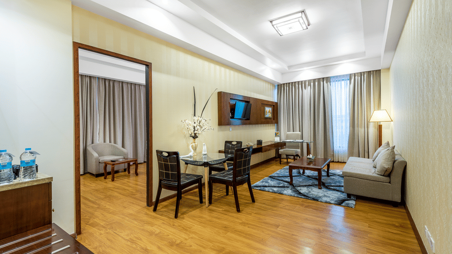 Modern hotel suite featuring a living area with a grey sofa, dining table, and wood flooring, with a doorway leading to a bedroom at Golden Tulip Kukas, Jaipur.