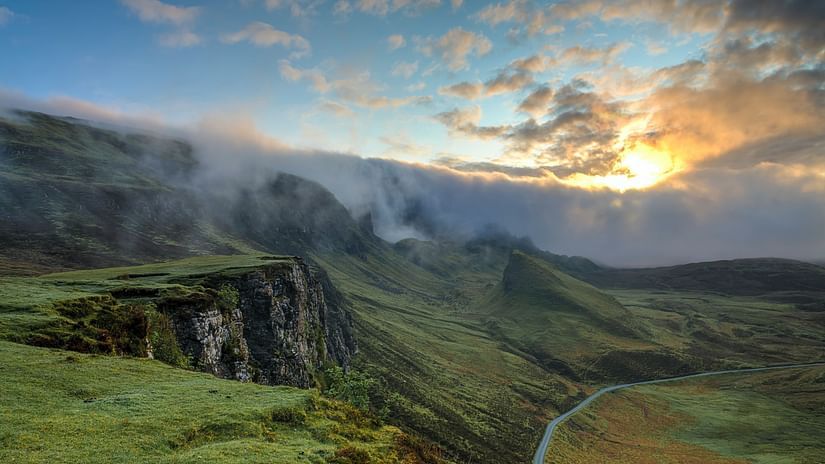 A dramatic landscape with fog rolling over green cliffs and valleys at sunrise or sunset, with a colourful sky