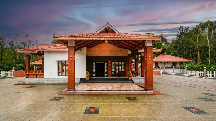 An eye-level shot of the main entrance to Bhuthanakadu Retreat, highlighting the wooden archways and the patterned tiled courtyard under a colorful sunset.