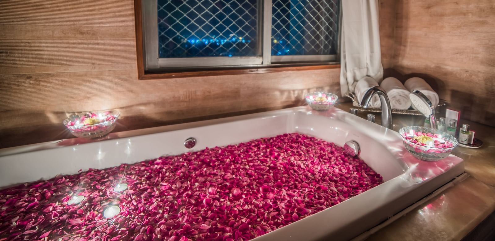 A romantic setting with a bathtub filled with rose petals and surrounded by candles, with a window overlooking the night sky A grand hotel suite with an opulent red and white decor, large bed, and seating area, reflecting a traditional style at Sitara Luxu