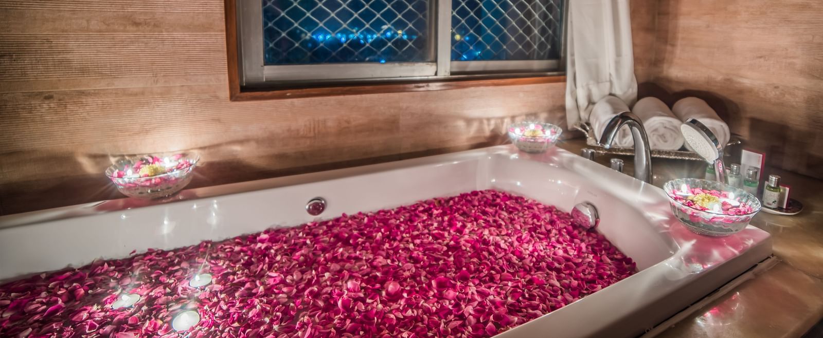 A romantic setting with a bathtub filled with rose petals and surrounded by candles, with a window overlooking the night sky A grand hotel suite with an opulent red and white decor, large bed, and seating area, reflecting a traditional style at Sitara Luxu