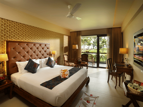 Lake View Deluxe Room at Uday Backwater Resort features a plush bed facing a wall-mounted TV, with a work desk beside it and a cosy seating nook by the window behind
