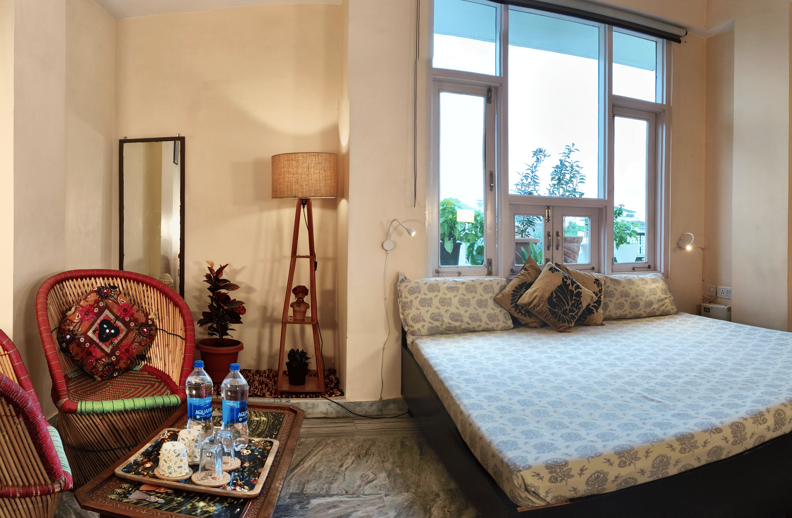 Bedroom with double bed, two chairs, a small table with bottles and tray, lamp and window view at Jaypore360 Homestay in Jaipur