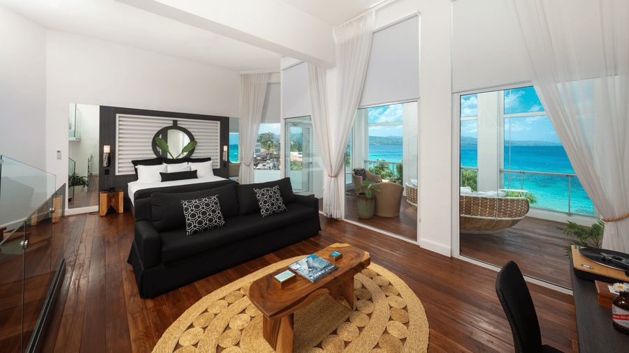 A wide-angle shot of a hotel room with a giant bed, a centre table, and a balcony offering views of the seaA peak into our room in Montego Bay with a bed placed in front of a wall mounted television and a couch for relaxation - S Hotel Montego Bay, Jamaica
