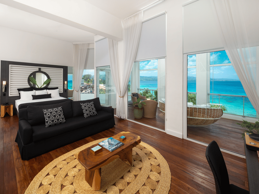 A wide-angle shot of a hotel room with a giant bed, a centre table, and a balcony offering views of the seaA peak into our room in Montego Bay with a bed placed in front of a wall mounted television and a couch for relaxation - S Hotel Montego Bay, Jamaica